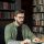 A man sits at a wooden table typing on a typewriter in a library filled with bookshelves
