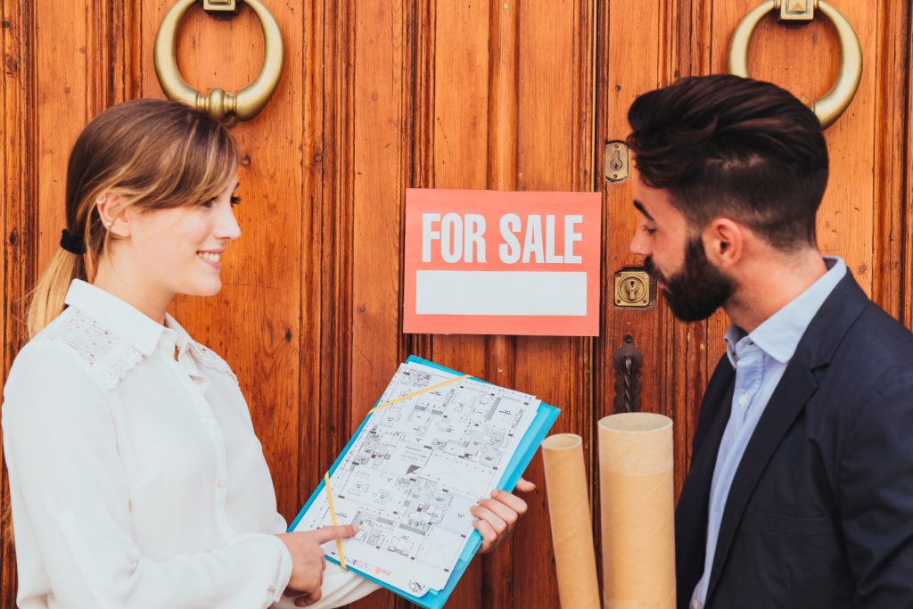 A man and woman stand together in front of a door displaying a "For Sale" sign
