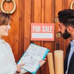 A man and woman stand together in front of a door displaying a "For Sale" sign